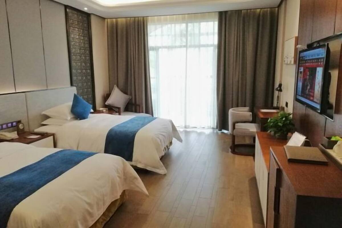 A hotel room with two beds, a desk, a chair, a TV, and a large window with curtains.