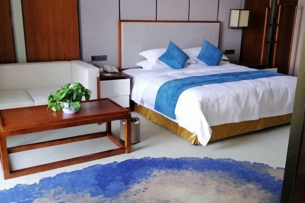 A hotel room with a bed, a wooden nightstand, a white sofa, a small table with a plant, and a blue and white patterned rug.