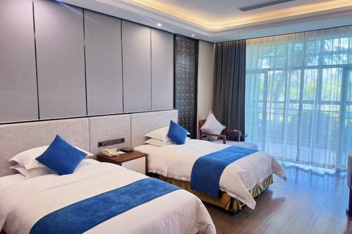 A hotel room with two beds, a large window with curtains, a nightstand, and a chair.