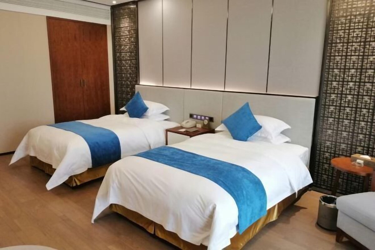 A hotel room with two beds, each with white and blue bedding, a wooden floor, a small bedside table with a phone, and a decorative wall panel.