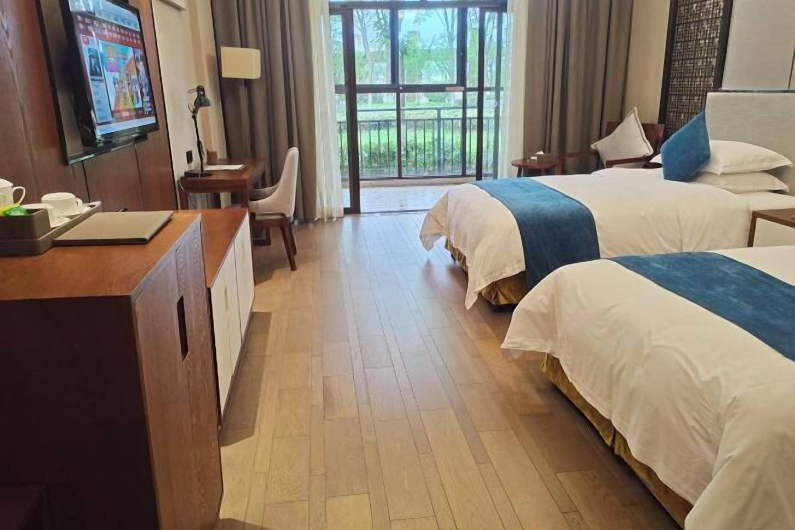 Habitación de hotel con dos camas, un escritorio con ordenador, un televisor y un ventanal con vistas a zonas verdes.