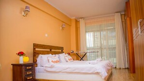 A bedroom with a wooden bed, a nightstand, a lamp, and a window with curtains. - Impeccable Family Friendly Apartment in Nairobi (Nairobi)