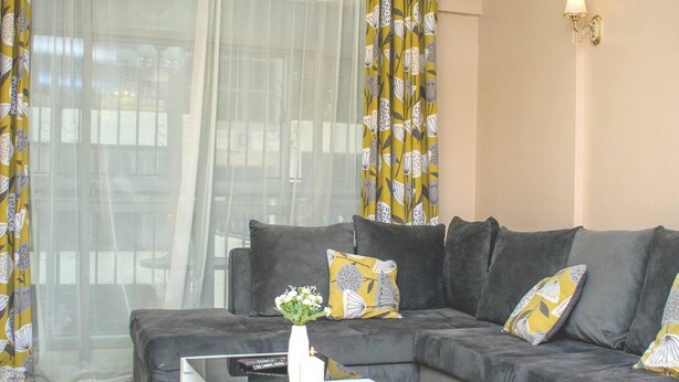 A living room with a grey sectional sofa, a white coffee table, and yellow patterned curtains. - Impeccable Family Friendly Apartment in Nairobi (Nairobi)