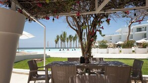 An outdoor seating area with wicker chairs and a table, overlooking a beach with palm trees and a pool.