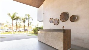 A reception desk with a marble counter and a wooden base, a wall-mounted plate, and a view of palm trees and parked cars in the background.
