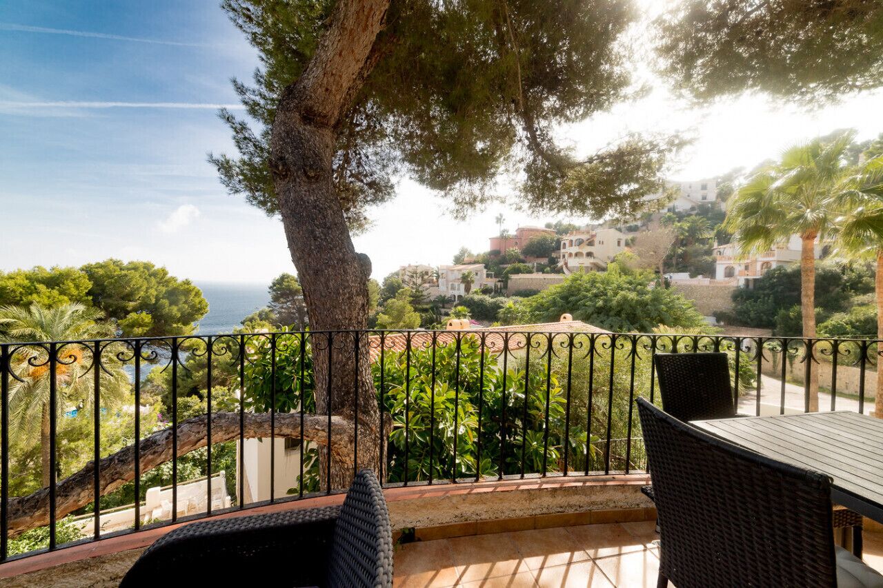 Ein Balkon mit Korbmöbeln, Blick auf eine Küstenstadt und ein Baum im Vordergrund.