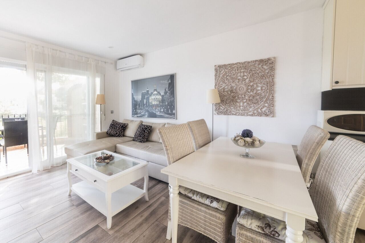 Una sala de estar moderna con sofá, mesa de centro y mesa de comedor. Hay un ventanal con cortinas translúcidas, aire acondicionado instalado en la pared y un cuadro con una vista urbana enmarcada.