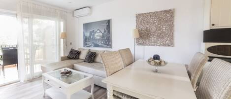 Una sala de estar moderna con sofá, mesa de centro y mesa de comedor. Hay un ventanal con cortinas translúcidas, aire acondicionado instalado en la pared y un cuadro con una vista urbana enmarcada.