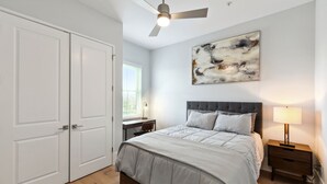 A bedroom with a bed, a desk, a chair, a nightstand, a ceiling fan, and a window.