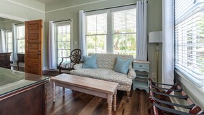 Une pièce avec un plancher en bois, un canapé agrémenté de coussins à motifs, une table, une chaise, une lampe et une fenêtre avec des stores.