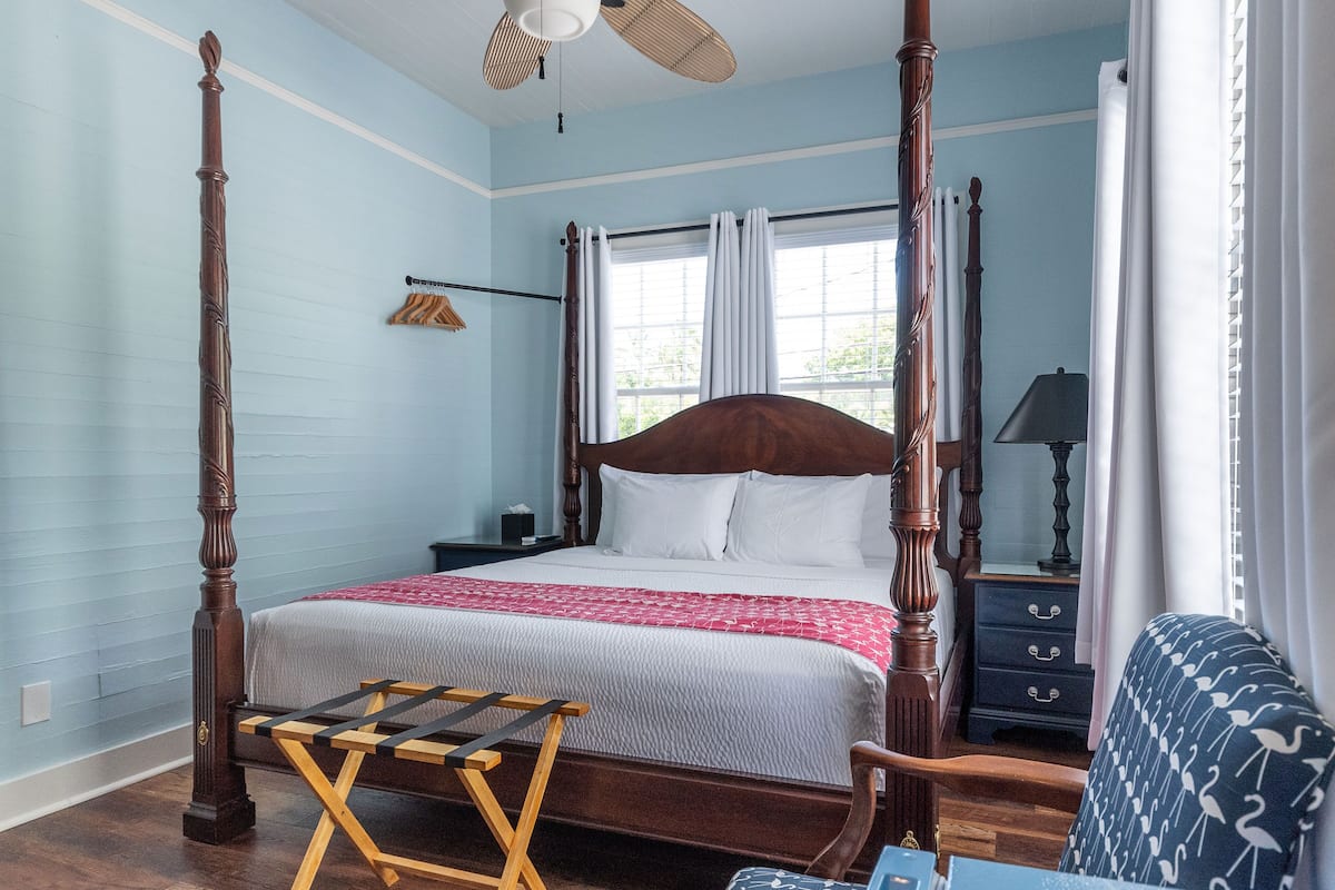 A four-poster bed with a wooden headboard, a wooden bench, and a patterned armchair in a room with light blue walls and a wooden floor.