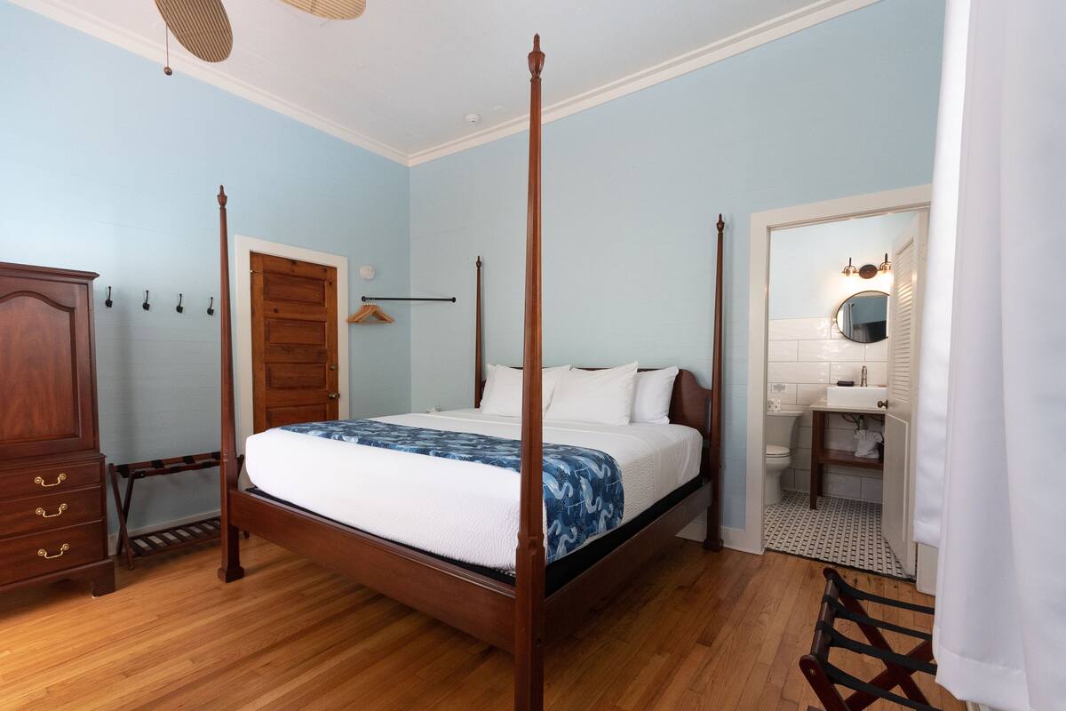 A four-poster bed with a white and blue patterned bedspread, a wooden wardrobe, a wooden chair, and a bathroom with a white sink and mirror.