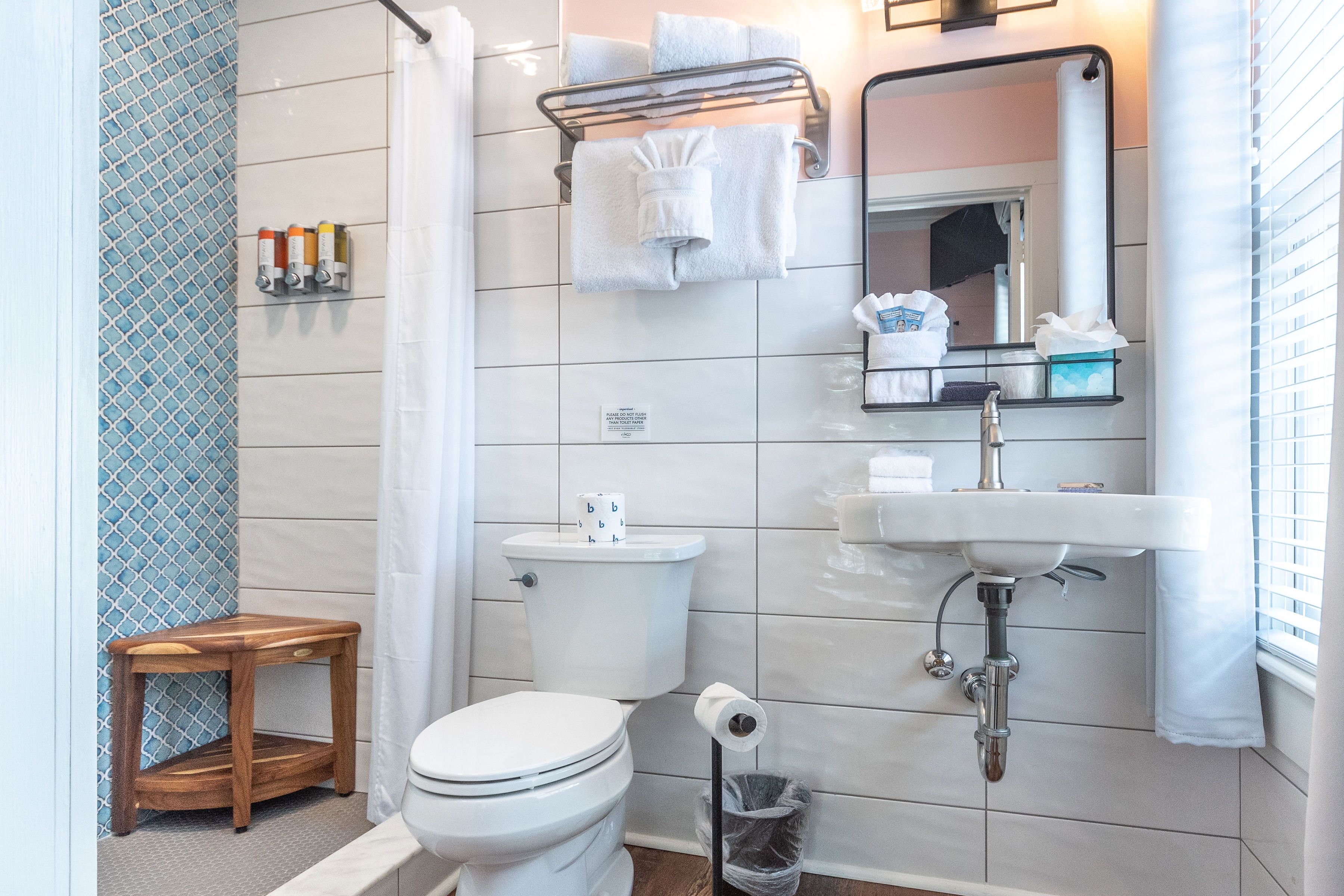 Et moderne badeværelse med toilet, vask og brusebad. Der er en lille træskammel og en skraldespand.