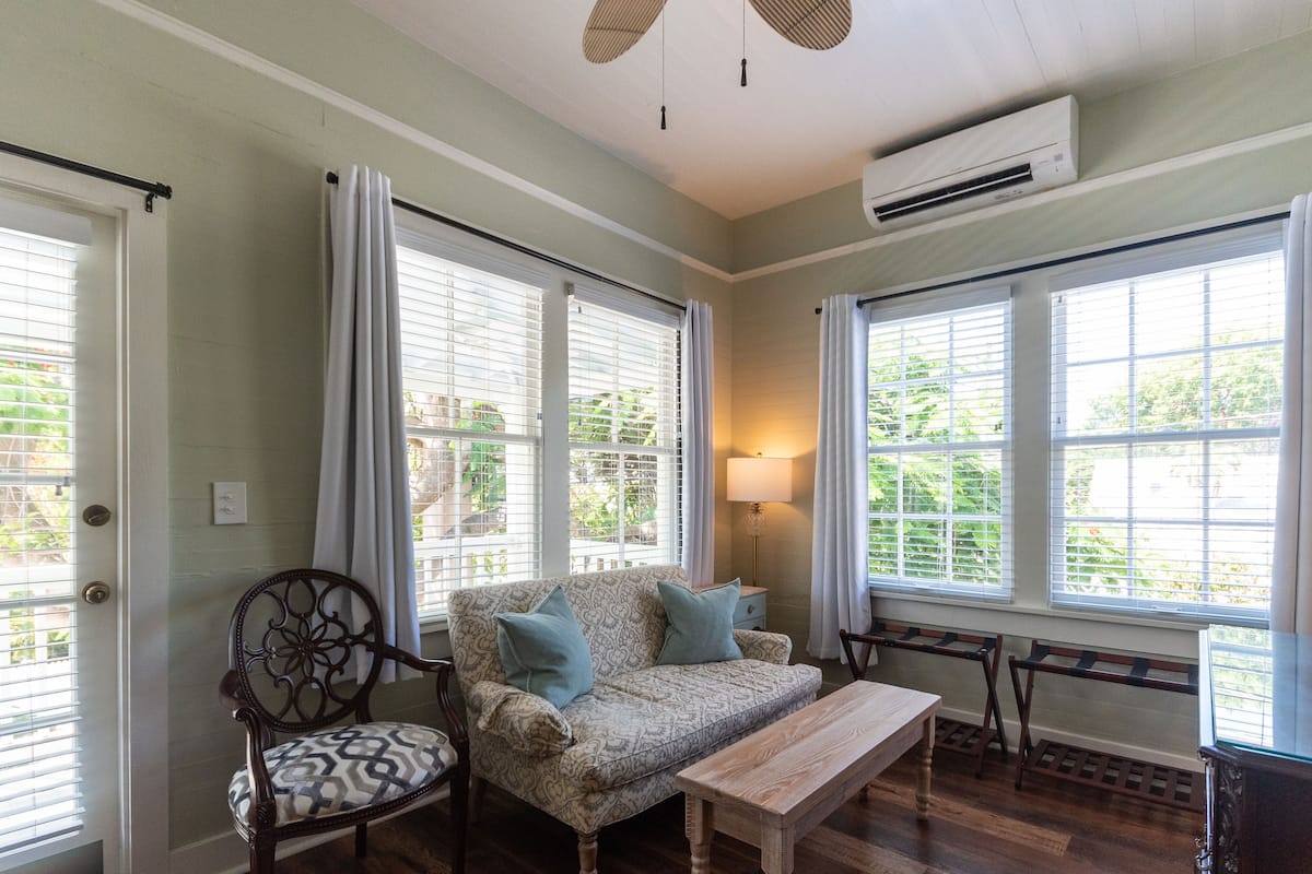 A room with a patterned sofa, a wooden bench, and a chair. There are large windows with blinds, a lamp, and an air conditioning unit.