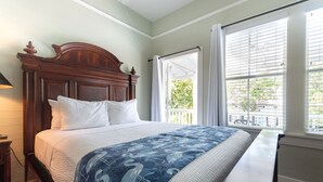 Une chambre à coucher avec une tête de lit en bois, un lit recouvert de linge de lit blanc, un plaid à motifs bleus et une vue par une fenêtre équipée de stores.