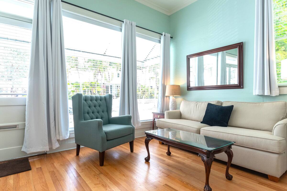 A living room with a teal armchair, a beige sofa, a glass coffee table, and a large window with curtains.