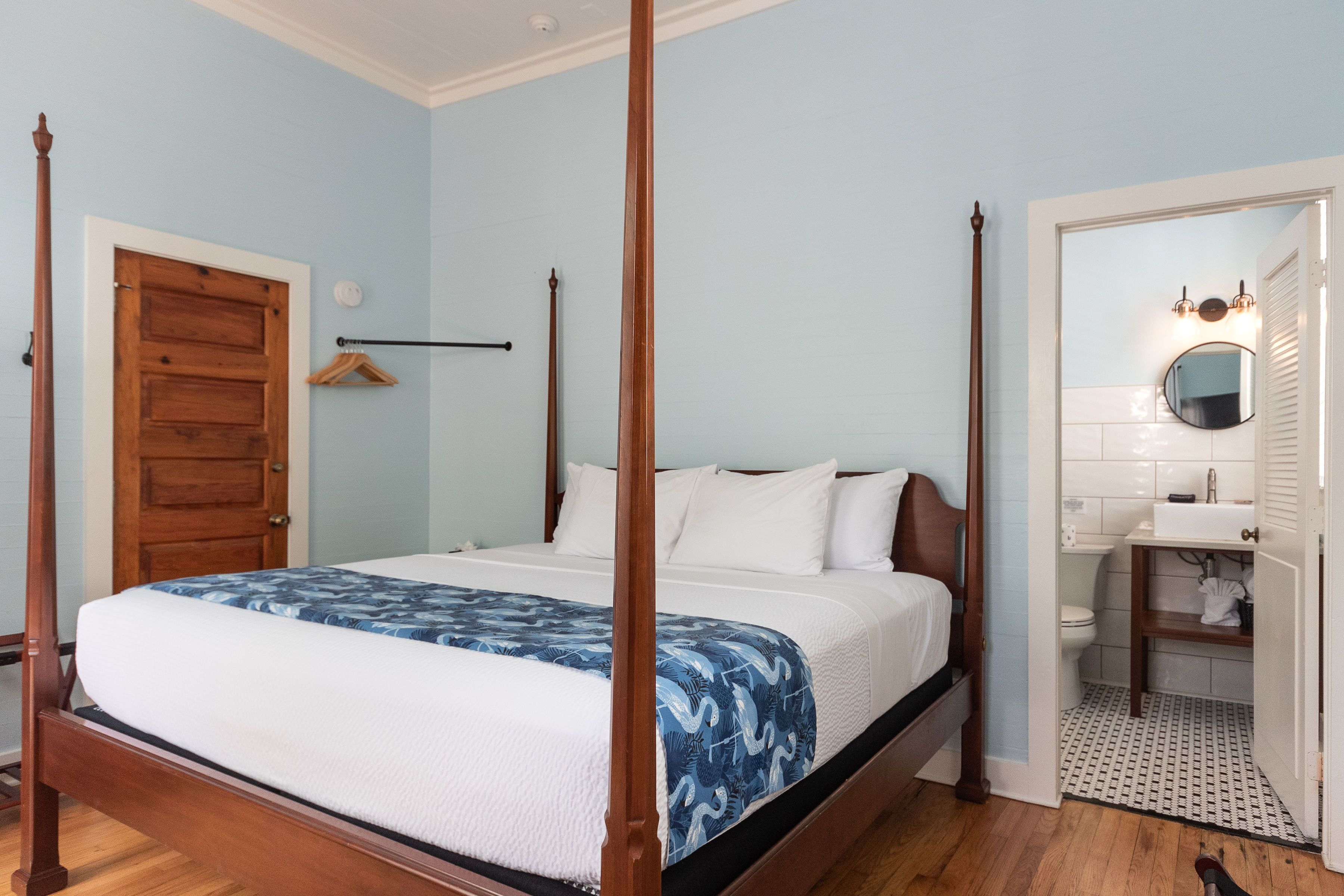 A four-poster bed with a white and blue patterned bedspread, a wooden door, a wooden wardrobe, and a bathroom with a white sink and mirror.
