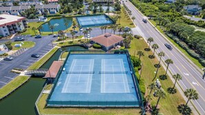 An aerial view of a residential area with two tennis courts, a parking lot, and a road with vehicles.