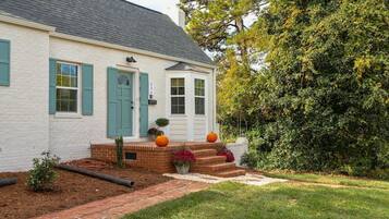 Una casa blanca con contraventanas verdes, un porche delantero de ladrillo y un pequeño jardín con calabazas.