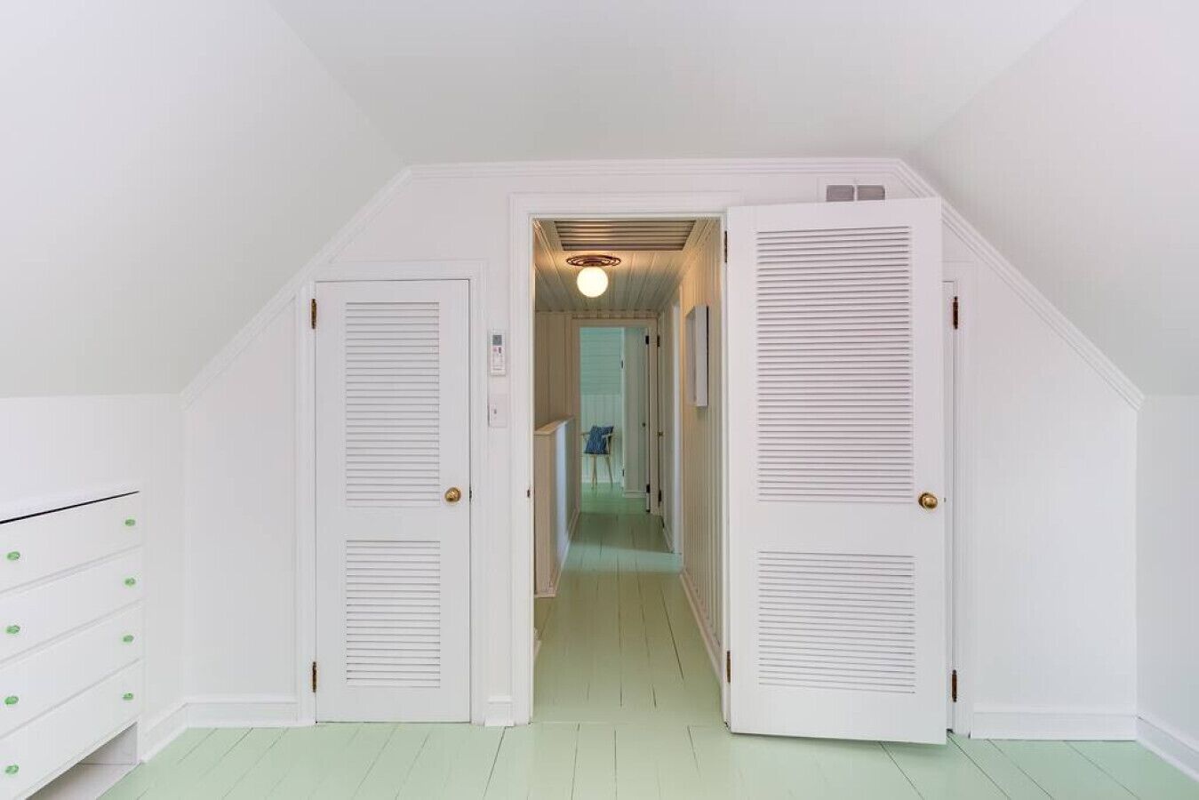 Un pasillo estrecho con puertas blancas y suelo verde claro, que conduce a una habitación con una lámpara en el techo.