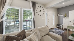 A living room with a large wall clock, a sofa with patterned cushions, a dining area with a table and chairs, and a view of the street through large windows.