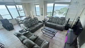 A modern living room with a sectional sofa, a coffee table, and a TV stand. Large windows provide a view of the ocean and palm trees. - Oceanfront dream condo 2/2 (Hollywood)