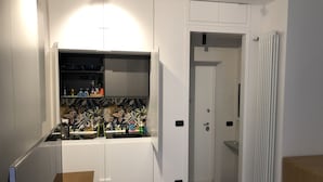 Une cuisine moderne avec des meubles blancs, un plan de travail sur lequel sont disposés divers ustensiles de cuisine, et une porte donnant sur une salle de bain.