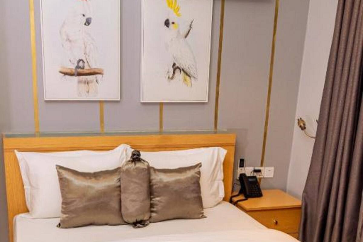 A neatly made bed with a brown headboard, a pillow, and a folded towel on the bed. There is a bedside table with a phone and a lamp. Two framed pictures are hung on the wall above the bed.