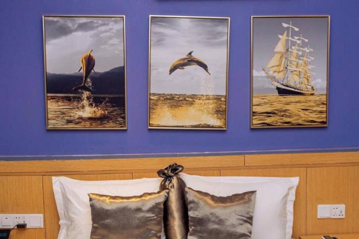 A hotel room with a bed, a wall-mounted phone, and three framed pictures depicting a sailboat, a dolphin, and a landscape.