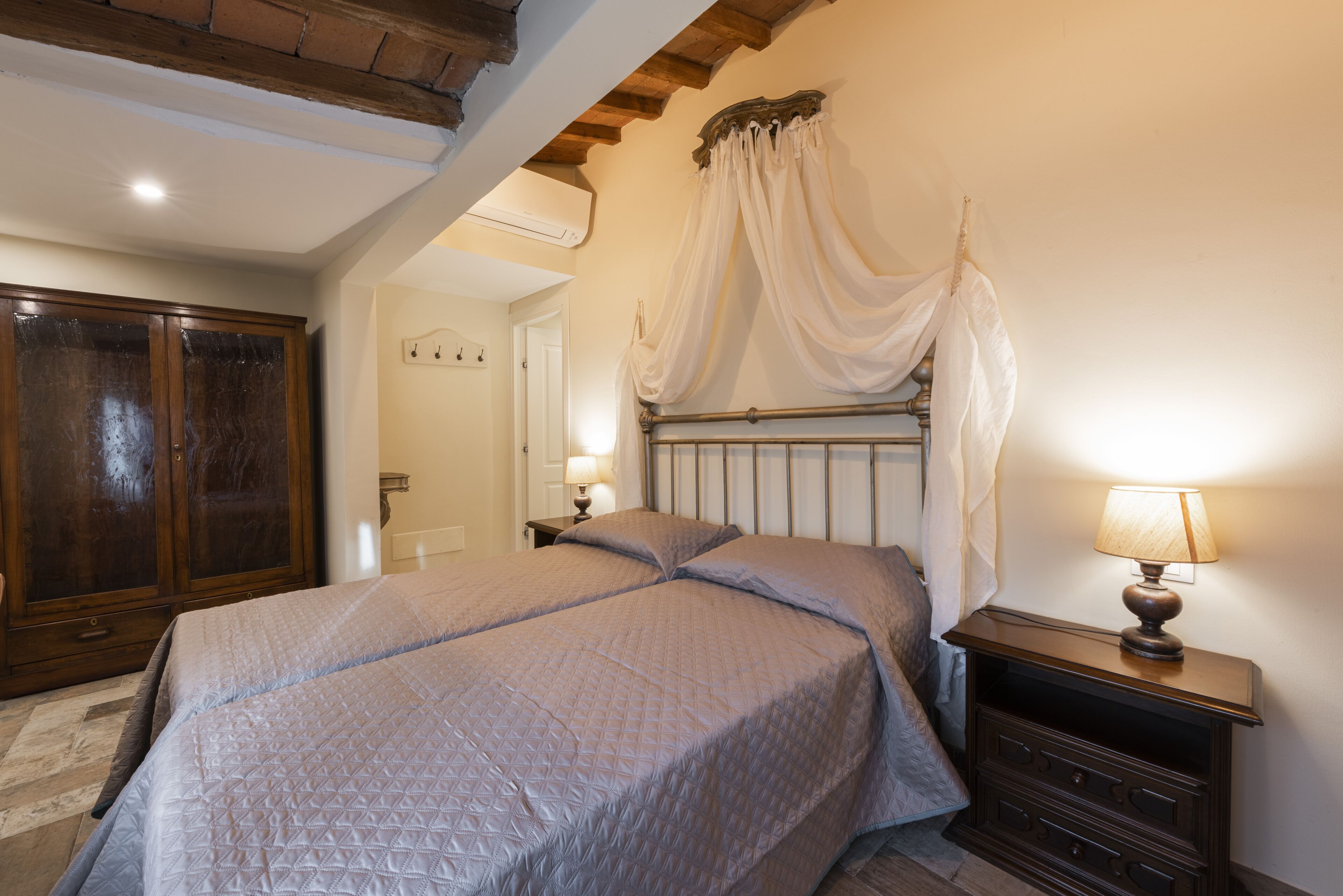 A bedroom with a large bed, a nightstand with a lamp, a wooden wardrobe, and a canopy bed.