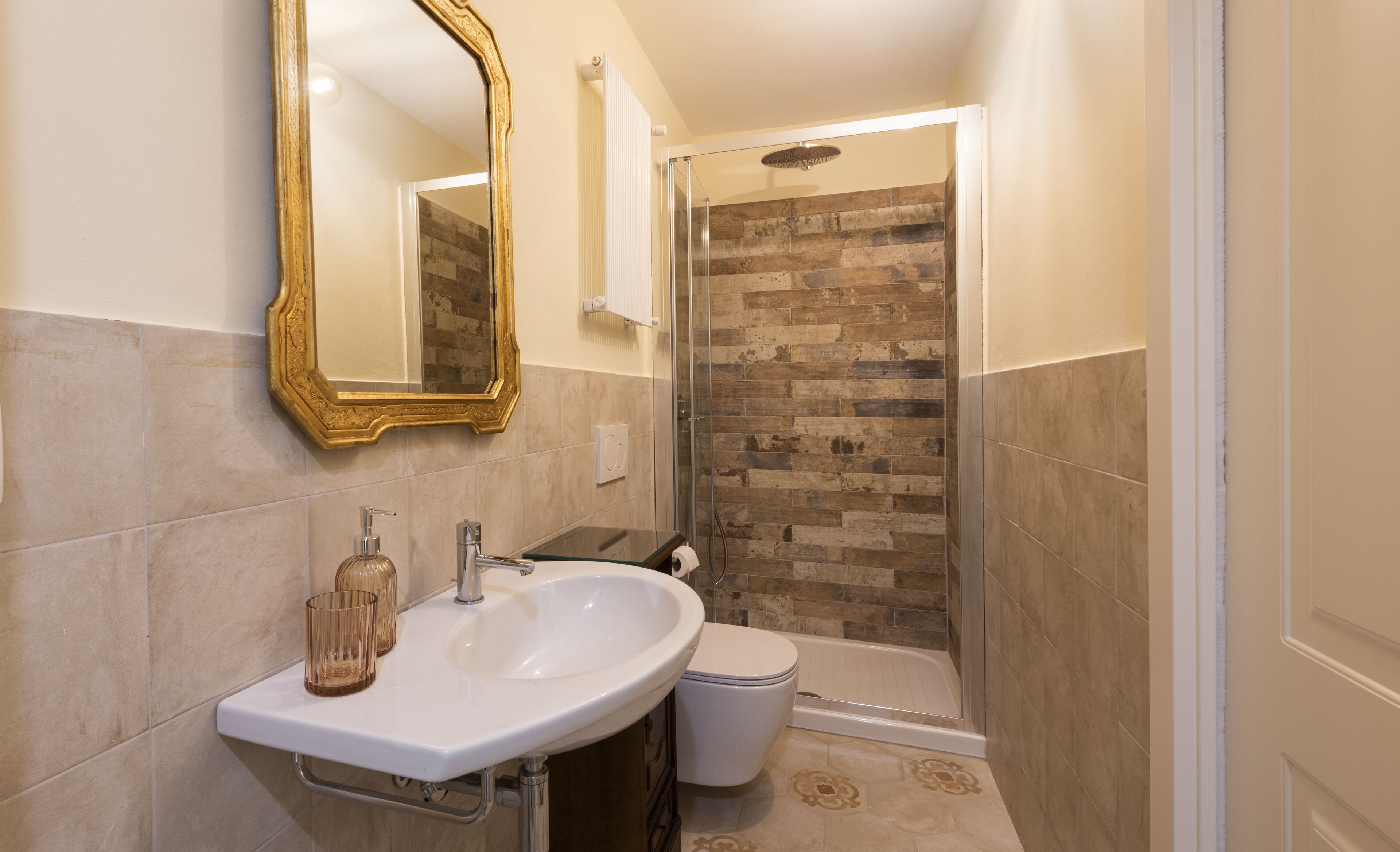 Un baño con lavamanos blanco, espejo con marco dorado, cabina de ducha de vidrio y retrete.