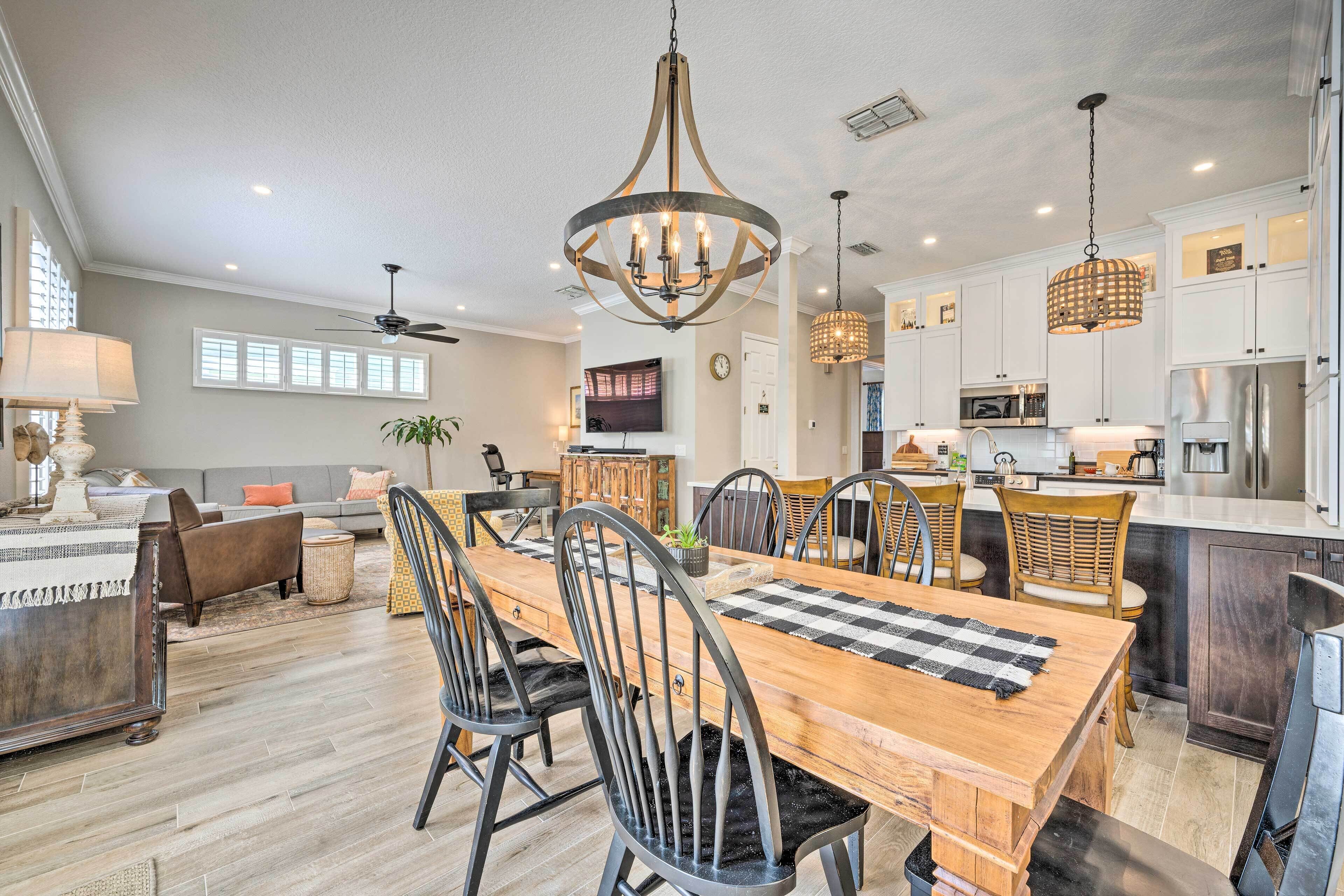 A spacious dining area with a wooden table and chairs, a chandelier, and a kitchen in the background.
