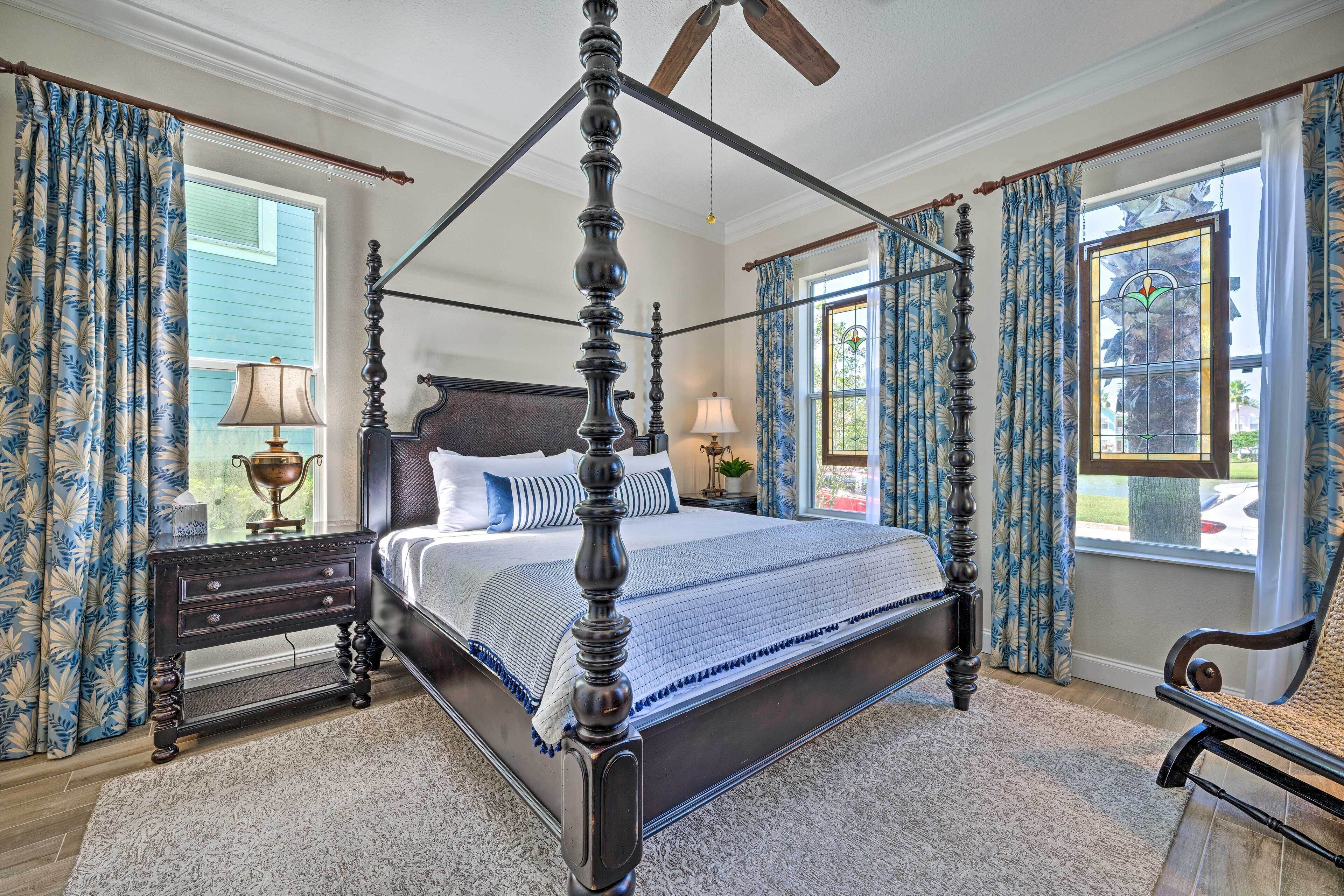 Un lit à baldaquin au revêtement foncé, agrémenté de deux tables de chevet assorties et d’un fauteuil, dans une pièce dotée de grandes fenêtres et de rideaux bleus.