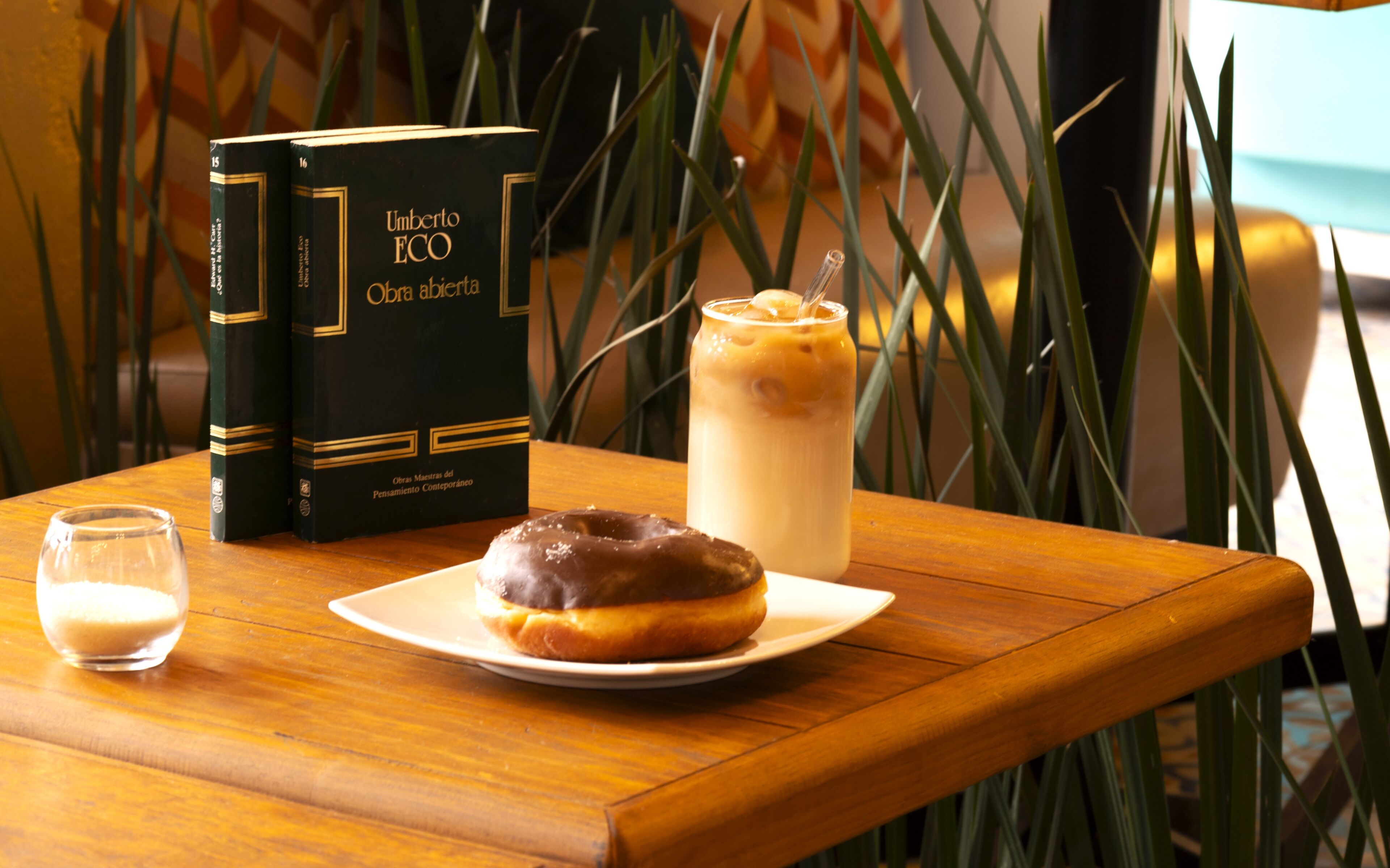 Una mesa con un libro, un donut y un vaso de café helado.