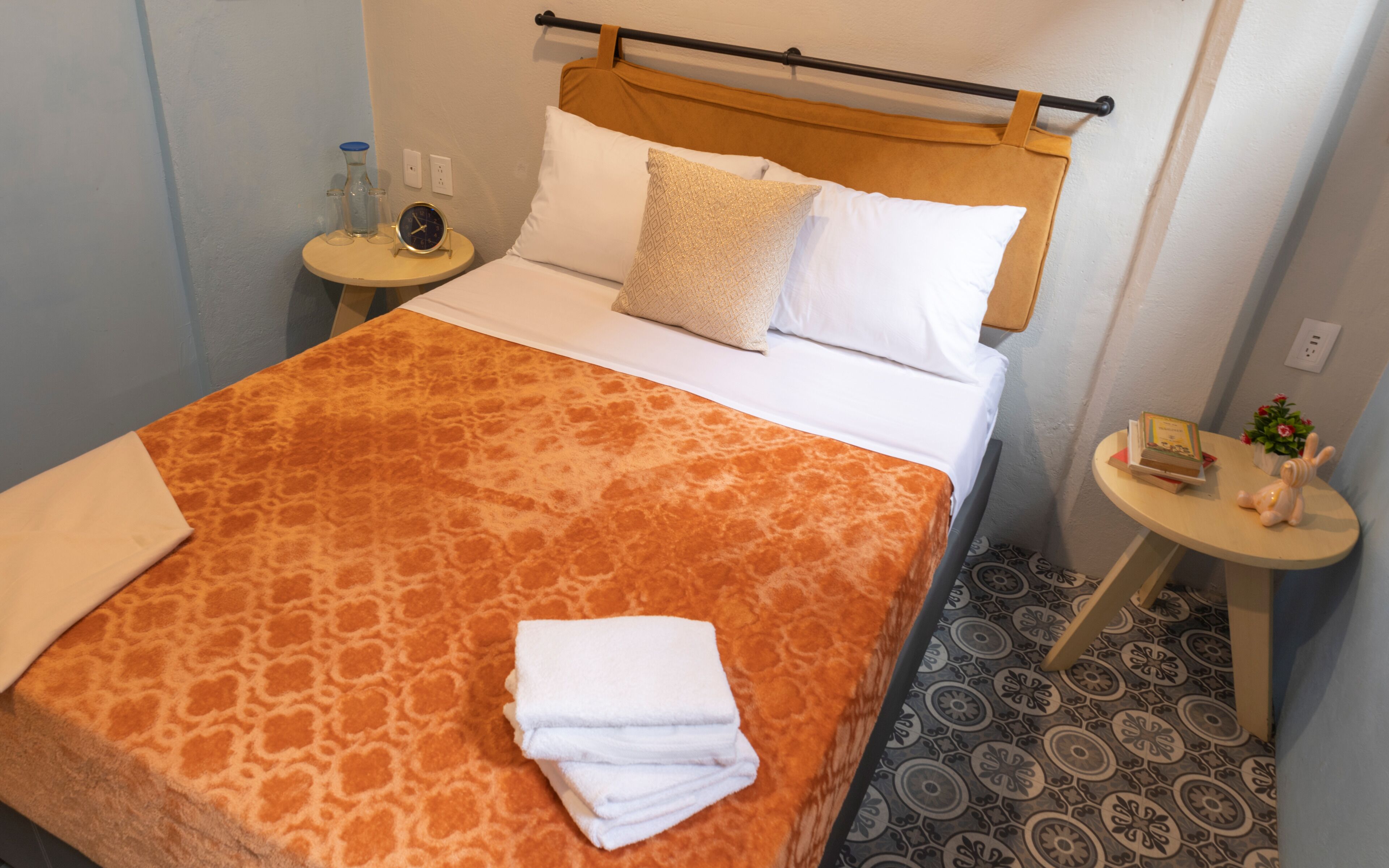 A neatly made bed with a patterned bedspread, a folded towel, and a bedside table with a clock and a small plant.