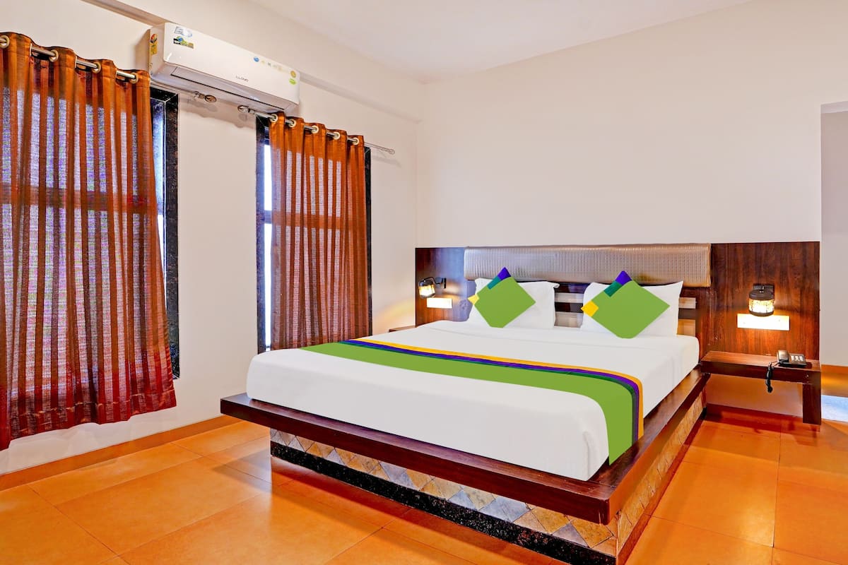 Quarto de hotel com cama, cortinas, ar condicionado, mesa de cabeceira com luminária e cabeceira de madeira.