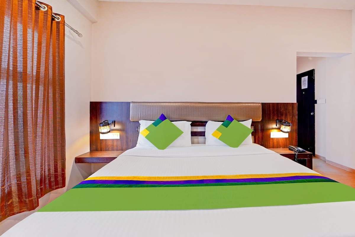 Quarto de hotel com uma cama grande, duas mesinhas de cabeceira, um criado-mudo e cabeceira de madeira, além de uma luminária fixa na parede.