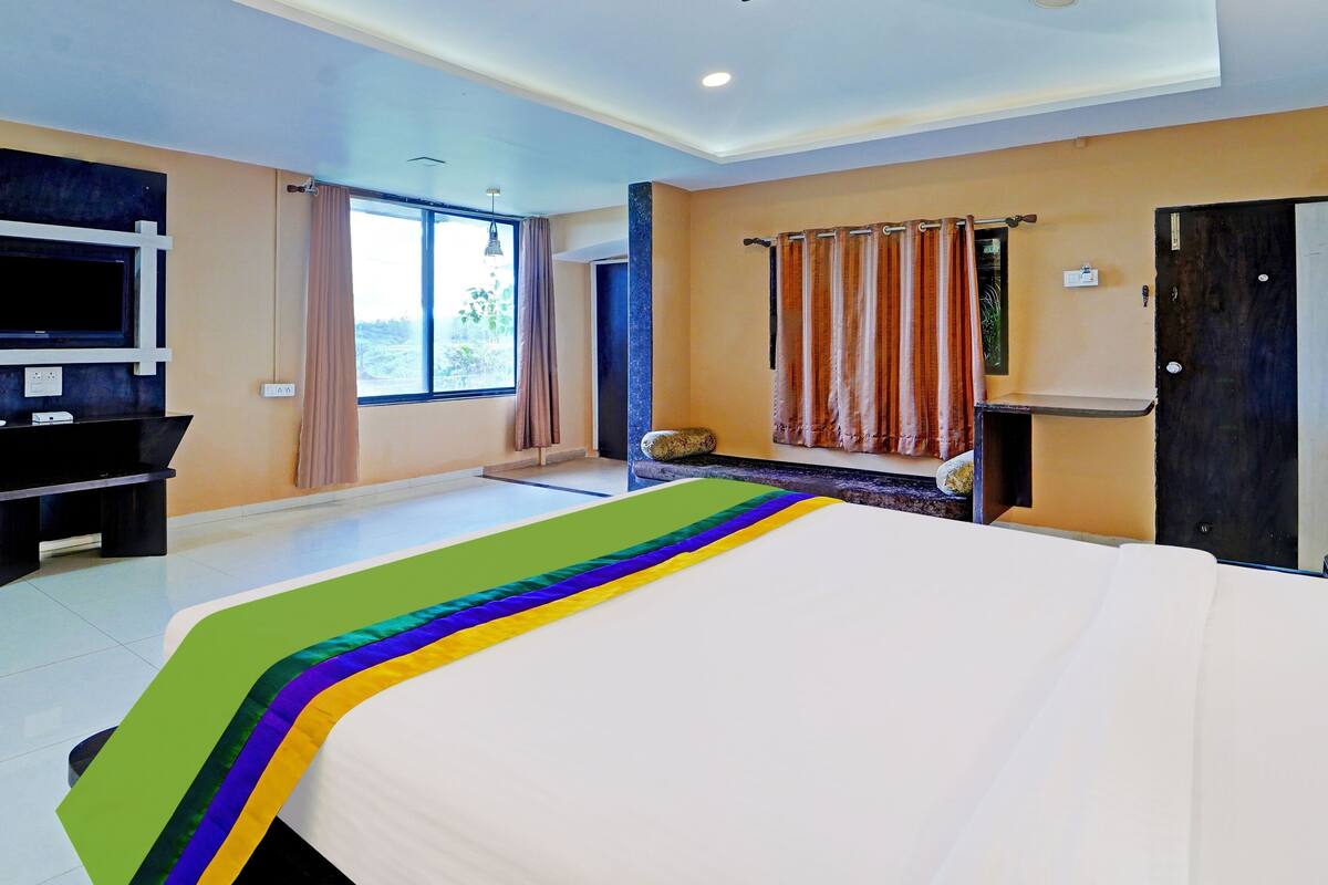 Quarto de hotel com cama, televisão, escrivaninha e janela com cortinas.