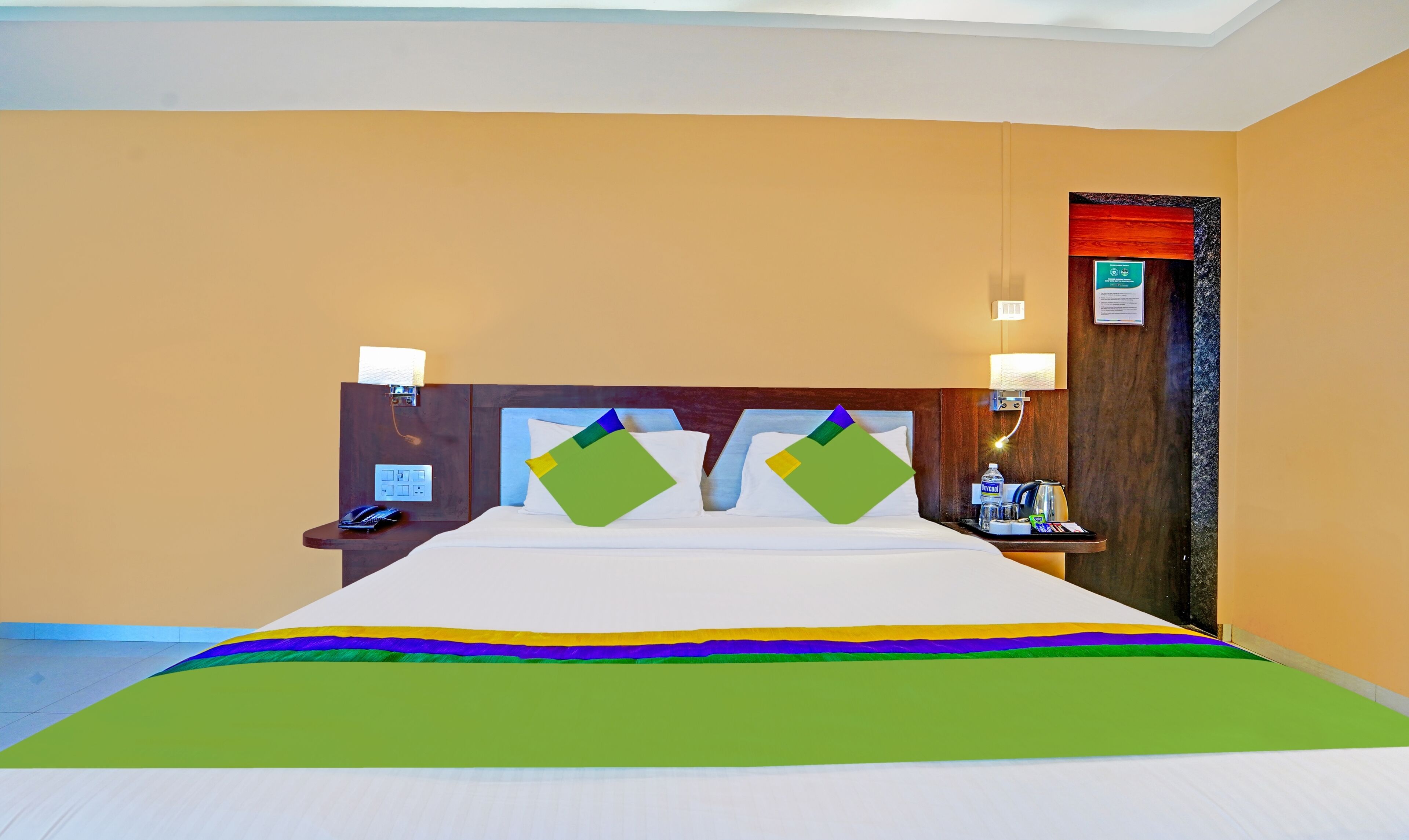 Habitación de hotel con una cama grande, dos lámparas de noche, una mesita de noche con un teléfono y una pequeña mesa con una botella de agua y una lámpara.