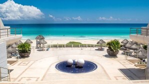 Et resort pool med et cirkulært vandelement, omgivet af liggestole og potteplanter, med udsigt over en strand og en klar blå himmel.
