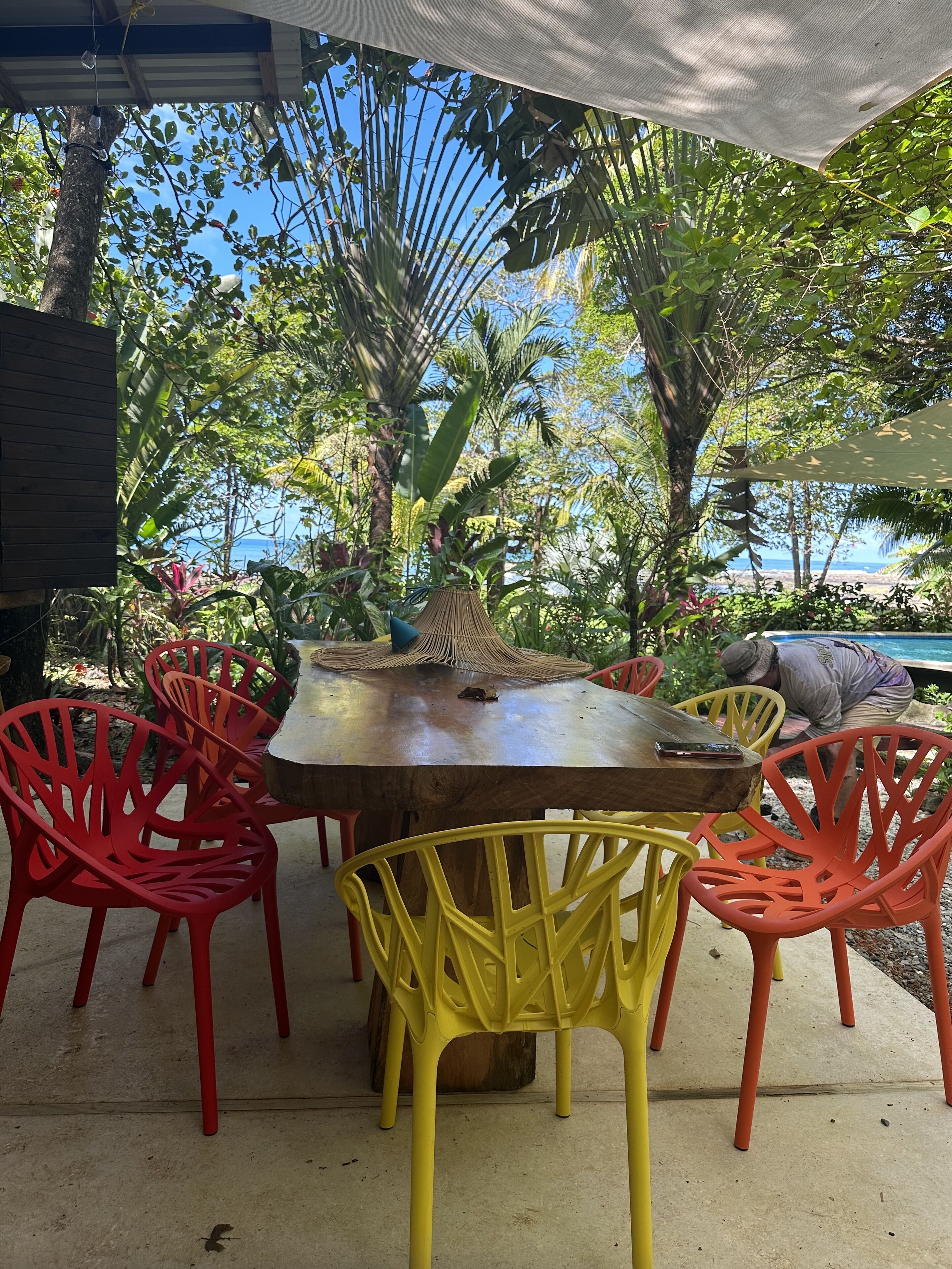 An outdoor dining area with a wooden table and colorful chairs, surrounded by lush greenery and a view of the sea.