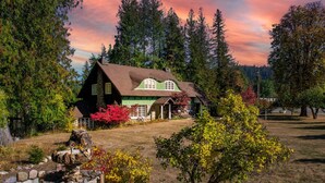 A rustic cabin with a stone foundation, a green roof, and a chimney. It is surrounded by a well-maintained lawn and trees. - The Kirtland Cutter Rock House - Metaline Falls (Metaline Falls)