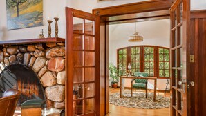A stone fireplace with a leather chair, a wooden door leading to a room with a green chair and a table, and a painting on the wall. - The Kirtland Cutter Rock House - Metaline Falls (Metaline Falls)
