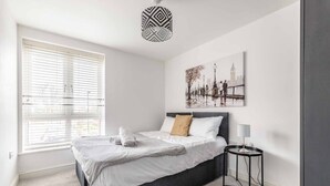 Ein modernes Schlafzimmer mit großem Fenster, einem Bett mit weißer Bettwäsche, einem schwarzen Kopfteil, einem Nachttisch mit Lampe und einem Stadtbild an der Wand.