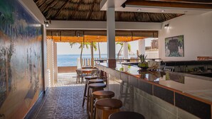 Un bar situé en bord de mer, avec un toit de chaume, des tabourets en bois et une vue sur l’océan.