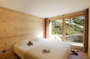 A bedroom with a wooden wall, a large window, a bed, a bedside table, a teddy bear, and slippers on the floor. - Chalet, view mountain, terrace, balcony, fireplace or stove, parking, tv, 162m², Les 2 Alpes (Les Deux Alpes)