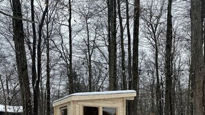 Una cabaña de madera pequeña con puerta de vidrio, rodeada de terreno y árboles cubiertos de nieve.