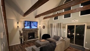 Una sala de estar con chimenea de piedra, un televisor de pantalla plana grande, un sofá gris, vigas de madera y un ventanal corredero que da a un espacio exterior.