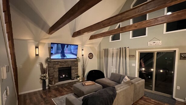 Una sala de estar con chimenea de piedra, un televisor de pantalla plana grande, un sofá gris, vigas de madera y un ventanal corredero que da a un espacio exterior.