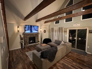 A living room with a stone fireplace, a large flat-screen TV, a grey sofa, wooden beams, and a sliding glass door leading to an outdoor area.